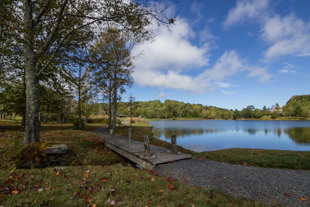 A tranquil scene featuring a wooden dock extending over a calm lake, surrounded by trees and distant rolling hills. The soft light suggests either early morning or late afternoon, enhancing the peaceful atmosphere. This image highlights the lakeside setting and the opportunities for fishing, picnicking, and nature observation right at the doorstep.