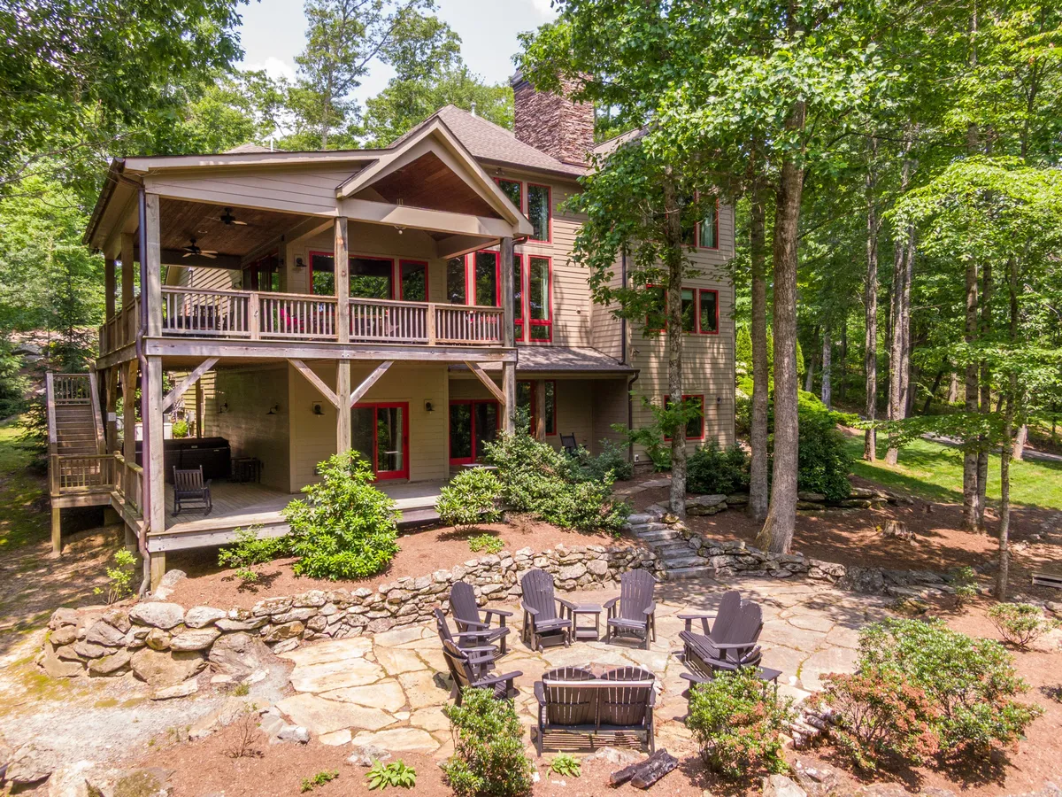 This image showcases the impressive exterior of Fern Ridge Lodge, with its expansive multi-level deck overlooking a stone fire pit area. The lodge is surrounded by lush green trees, creating a private, cozy mountain retreat. The setting suggests both relaxation and outdoor fun, with sunset views from the deck and the atmosphere of gathering around the fire pit amid nature.