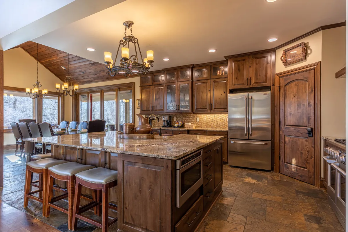 A spacious, open chef’s kitchen is featured here with a large kitchen island, gleaming granite countertops, double chandeliers, stainless steel appliances, and custom wood cabinetry. The design allows for both serious cooking and socializing around the island.