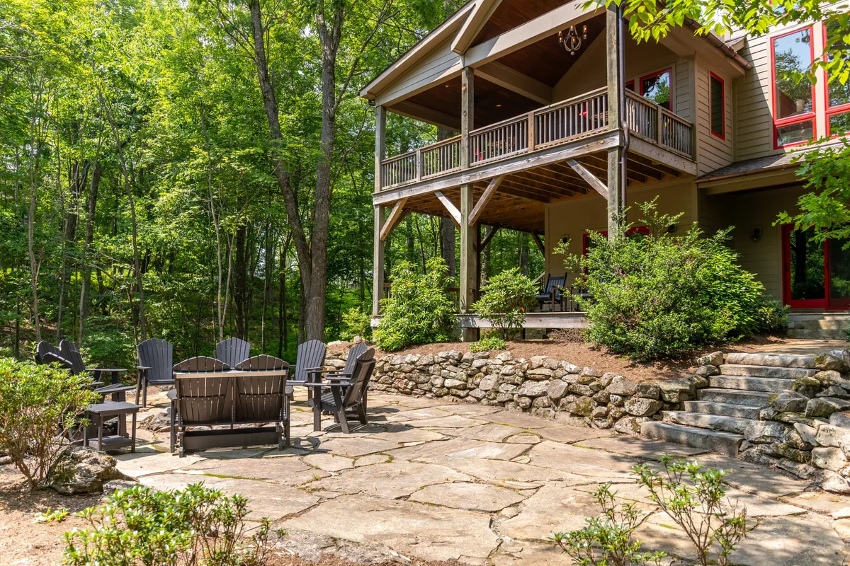 An inviting exterior shot showing a covered porch, a circular fire pit seating area with Adirondack chairs, a stone pathway, and lush greenery. The space is designed for communal gatherings around the fire and relaxation in the open air, with access to surrounding nature.