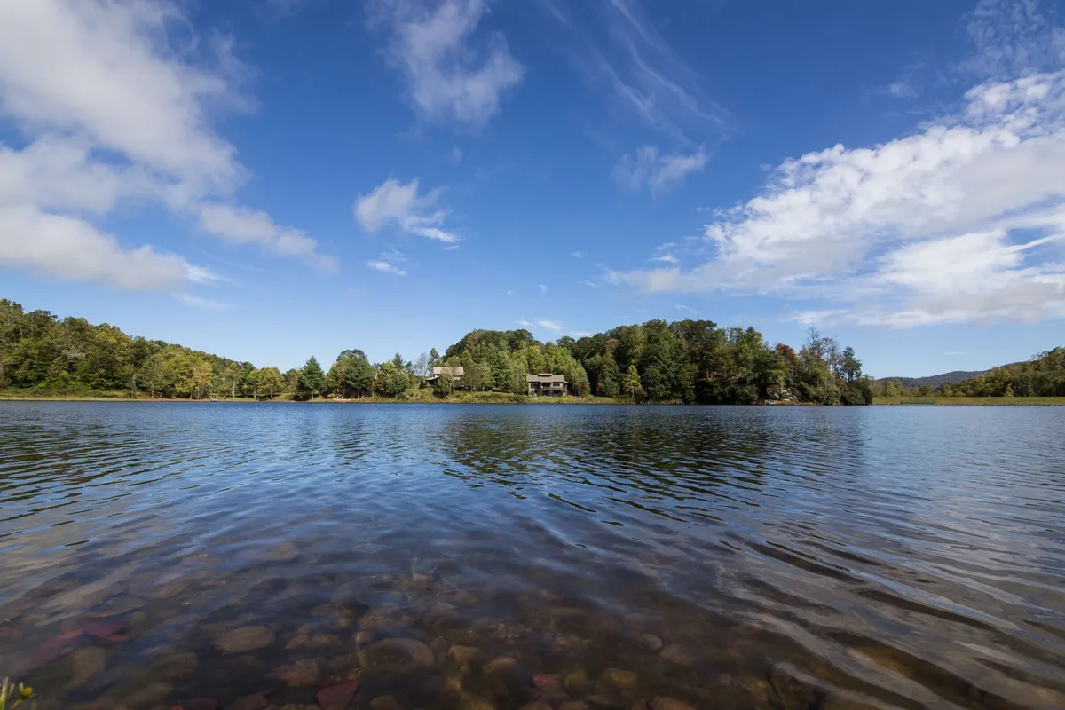 A serene view of a calm lake bordered by lush greenery, with a distant house visible across the water. The scene suggests opportunities for fishing, boating, nature walks, and lakeside picnics, complemented by a peaceful, unspoiled landscape ideal for stargazing or sunset viewing.