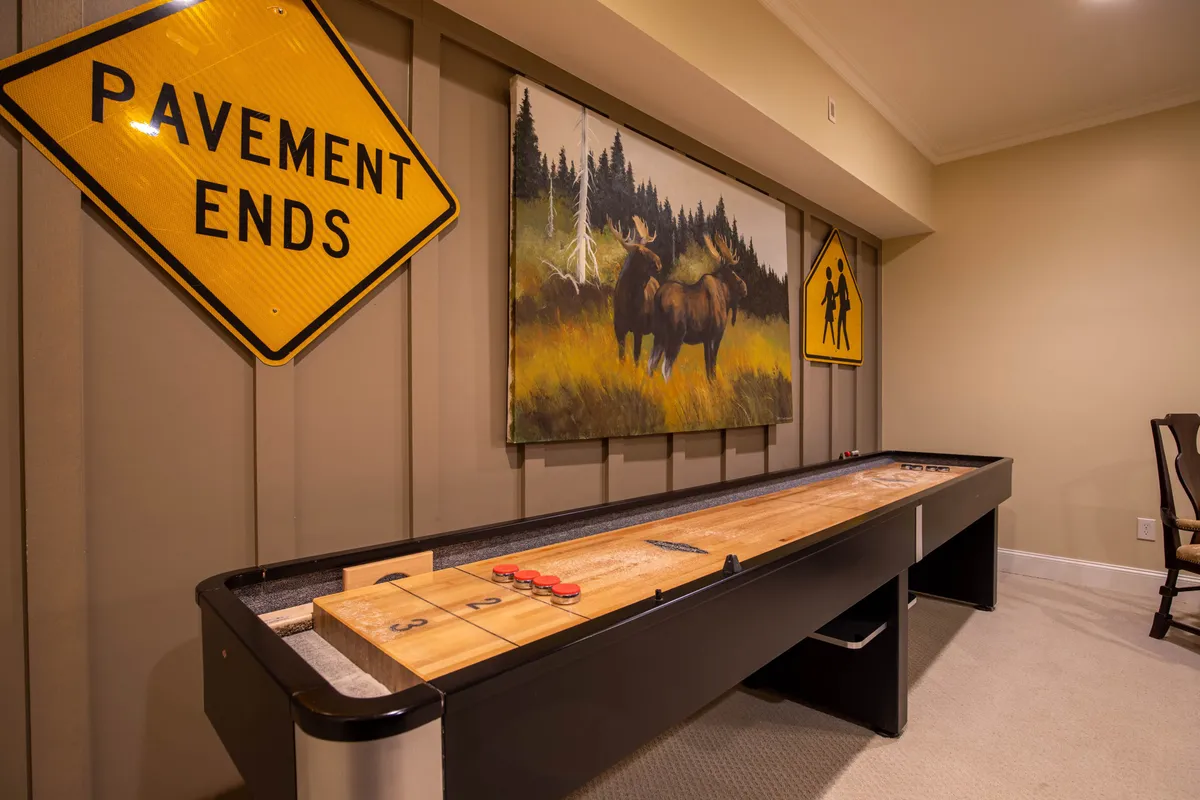 A vibrant interior view focused on a shuffleboard table, adorned with a playful moose painting and colorful warning signs, creating an inviting, lighthearted atmosphere. The space encourages friendly games and social interaction.