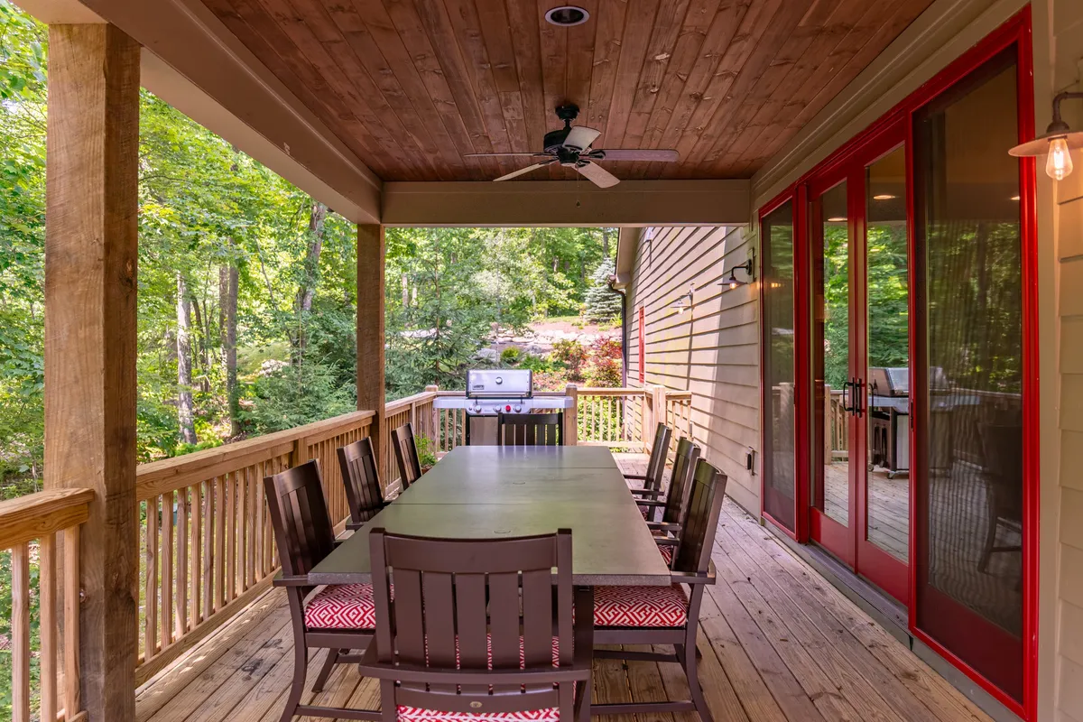 A spacious outdoor dining area features a large table, red accent doors, and an outdoor grill. The area is designed for hosting family meals, barbecues, and relaxed outdoor dining surrounded by greenery.