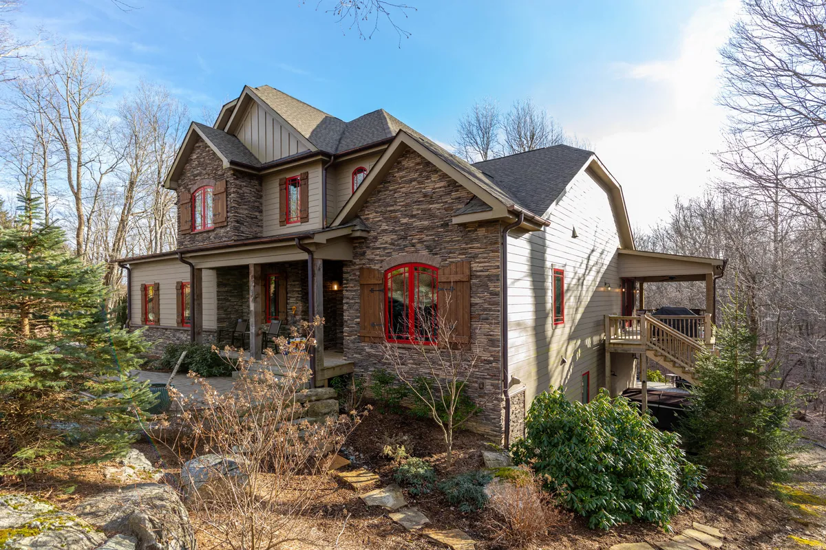 The image captures the home’s stone facade with red window frames, wooden shutters, and a beautifully landscaped garden. The porch area looks perfect for peaceful mornings or celebrating special occasions in the fresh mountain air.