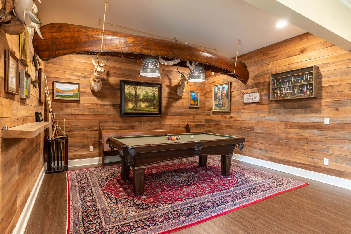 A cozy, recreational game room featuring a pool table beneath a unique wooden canoe lighting fixture, animal mounts on the wall, framed paintings, and a visible bar area in the background. The space is designed for friendly competition and social gatherings.
