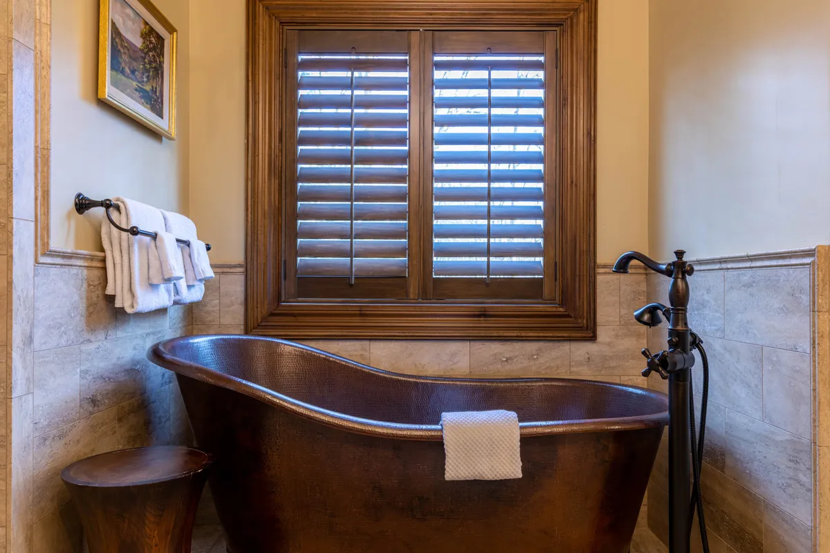 A spa-inspired bathroom with an opulent copper soaking tub, wooden window shutters, fresh towels on a rack, and a wooden stool. The space is tranquil and inviting, ideal for luxurious relaxation.