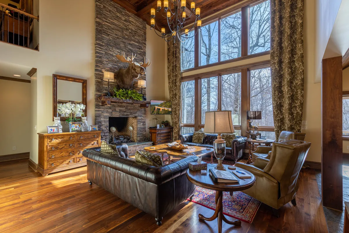This image showcases a grand great room with large windows that allow natural light to pour in, a prominent stone fireplace, a mounted moose head, leather sofas, and wooden furniture. The space radiates warmth and comfort, perfect for gatherings, relaxation, and fireside evenings.