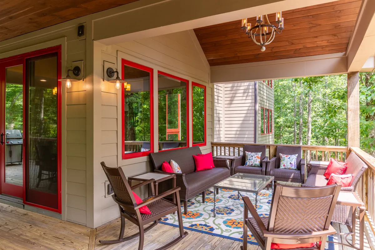 A cozy outdoor seating area with comfortable chairs, decorative pillows, a colorful rug, a glass coffee table, and a chandelier overhead. The space is surrounded by nature, inviting guests to relax outdoors in any season.