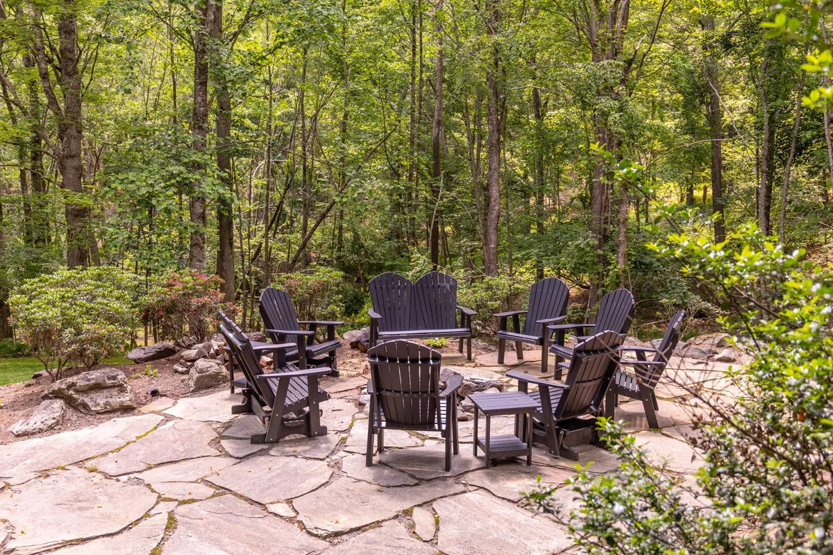 A circular arrangement of Adirondack chairs sits on a stone patio surrounded by tall trees, creating a focal point for gatherings. The natural setting offers a peaceful, forested backdrop, perfect for group activities and fireside conversations.