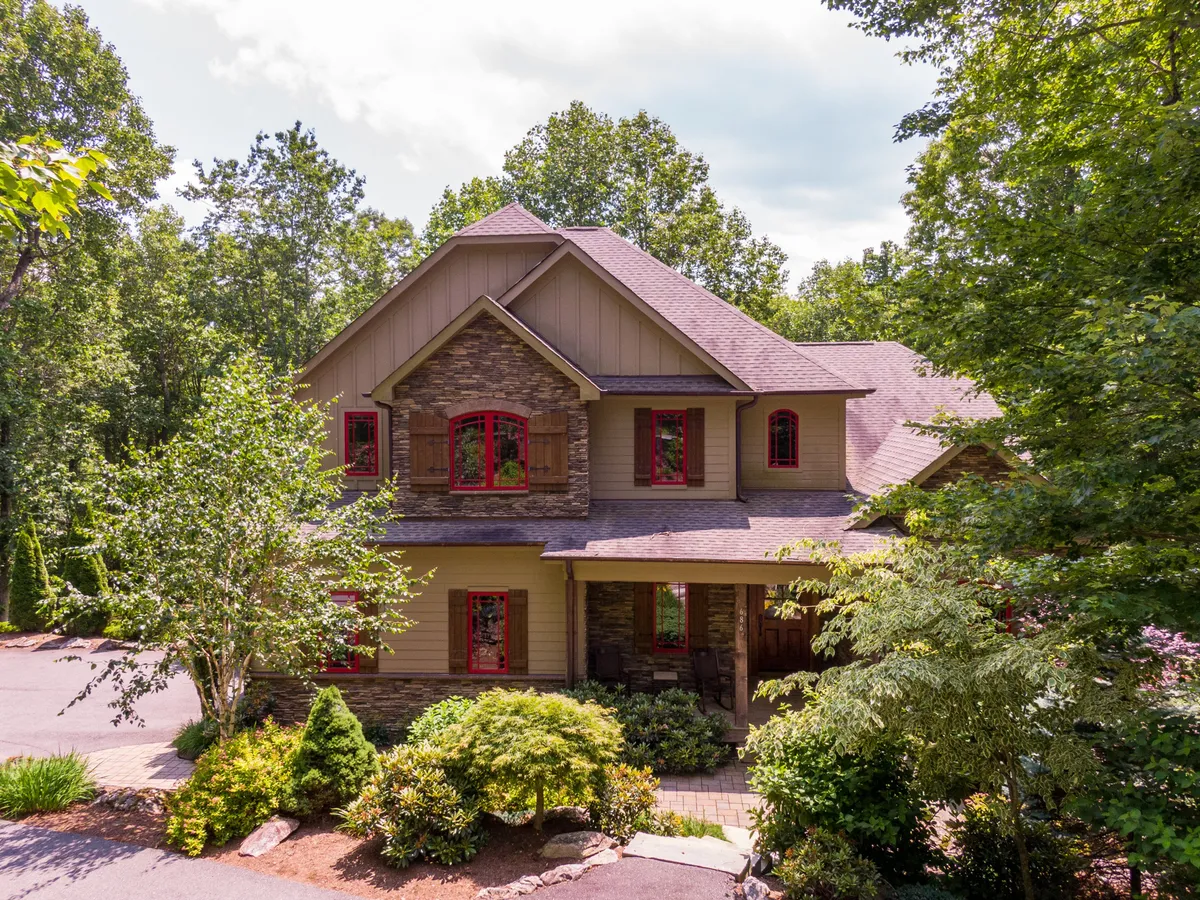 A broad view of the house's exterior featuring attractive stone details, signature red window frames, and lush landscaping. The home is nestled in greenery, hinting at year-round natural beauty and easy access to outdoor adventures.