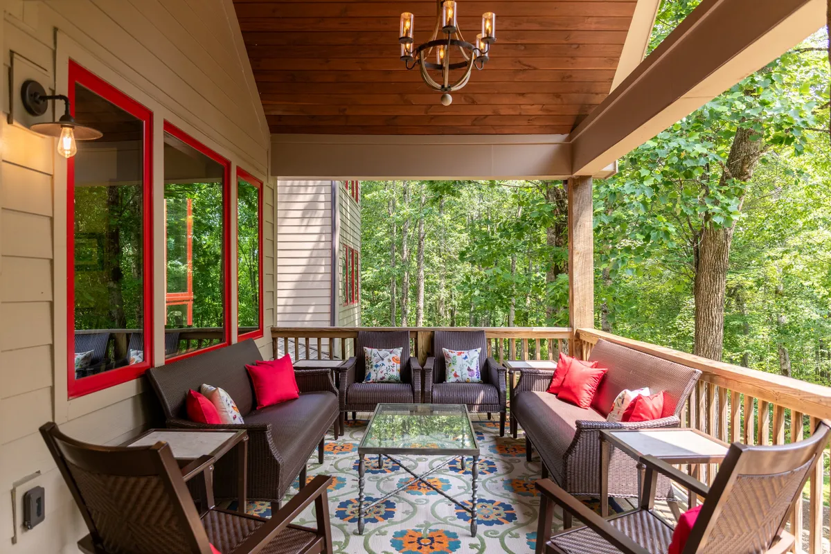 This exterior image features a comfortable seating area on a covered deck, complete with a colorful patterned rug and lush greenery visible beyond. The arrangement suggests a perfect spot for morning coffee, reading, or relaxing while immersed in nature.