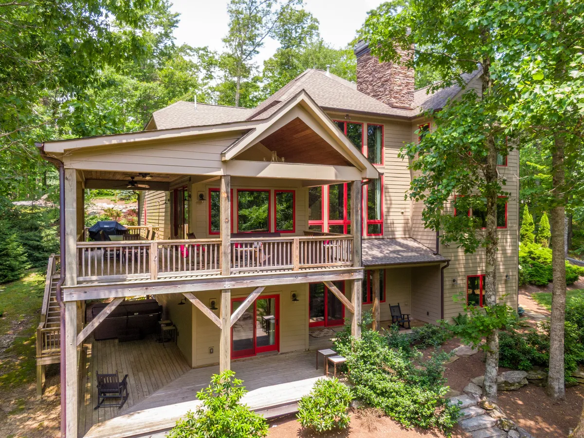 This image presents a spacious deck with red window frames, natural landscaping, and a stone chimney. The deck is surrounded by lush greenery and offers ample space for outdoor dining or relaxation while taking in the mountain views.