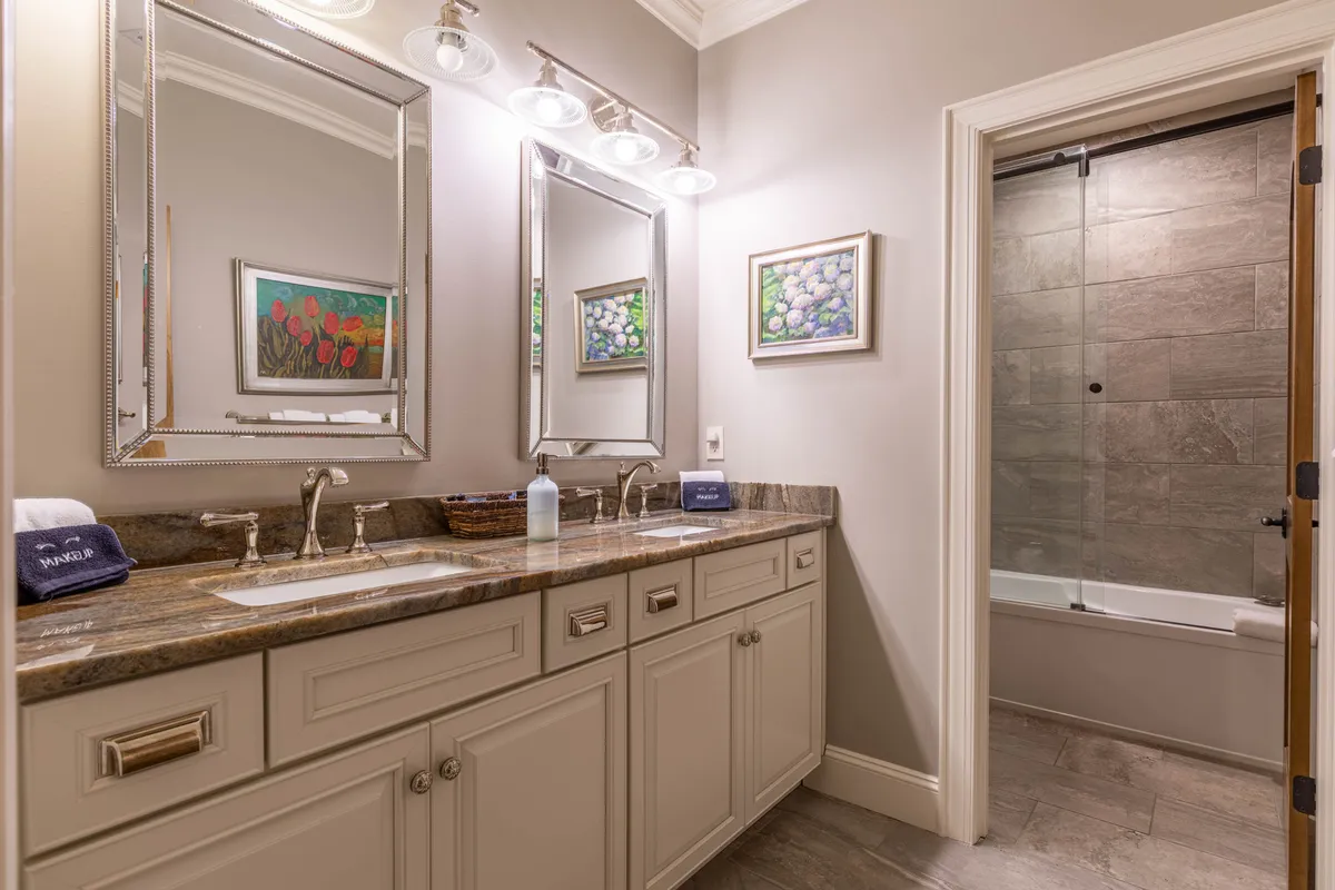 A spacious bathroom featuring a dual sink vanity, large mirrors, elegant tilework, framed artwork, and a glass shower door. The aesthetic is both serene and luxurious, evoking a spa-like retreat.